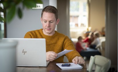 A man - Rob - carries out some admin at a computer, sat at his dining table. A calculate and notepad are at hand.