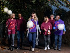 A group of six friends on the Walk of Light walk together at night time down a wide alleyway. They all wear Blood Cancer UK T shirts, and several of them carry globed lanterns held by long poles.