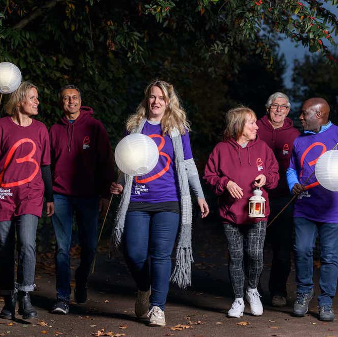 A group of six friends on the Walk of Light walk together at night time down a wide alleyway. They all wear Blood Cancer UK T shirts, and several of them carry globed lanterns held by long poles.