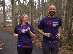 Two runners - a man and a woman - enjoy a run down a forest path. They both wear purple Blood Cancer UK t-shirts.
