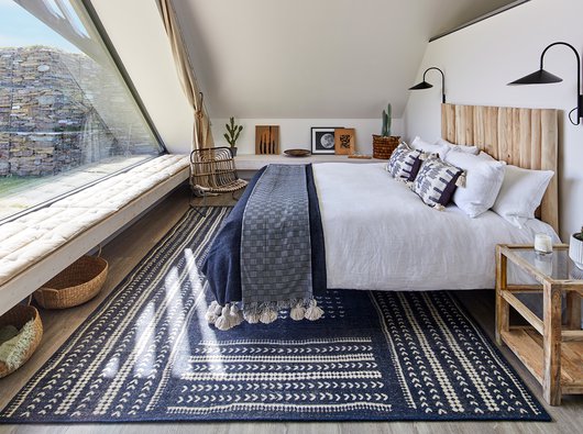 A well-dressed bedroom with double bed placed on a large navy rug, a slanted roof and wall-length windows make the space appear cosy and bright