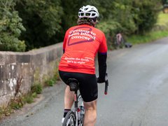 A cyclist heading down a country lane, wearing a Blood Cancer UK T shirt with "Cycling to beat blood cancer' on the back.