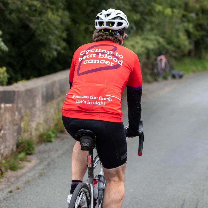 A cyclist heading down a country lane, wearing a Blood Cancer UK T shirt with "Cycling to beat blood cancer' on the back.