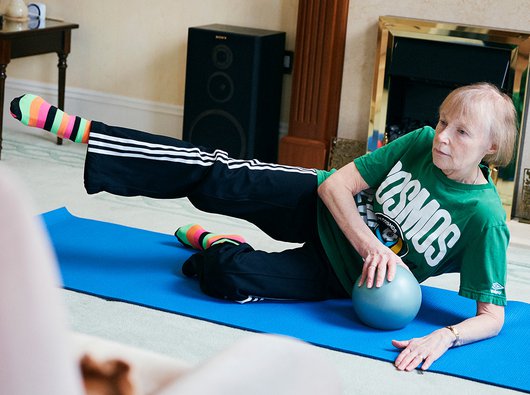 A woman in her 60s exercises in her living room on a yoga mat, using an exercise ball. She's wearing a green t-shirt with a logo and black jogging bottoms with three white accent stripes.
