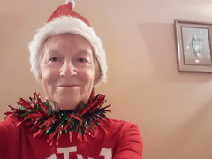 An older woman looks direct to camera, wearing a santa hat and tinsel around her neck. She's indoors, at home.