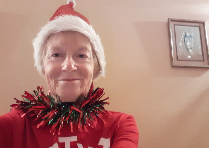 An older woman looks direct to camera, wearing a santa hat and tinsel around her neck. She's indoors, at home.