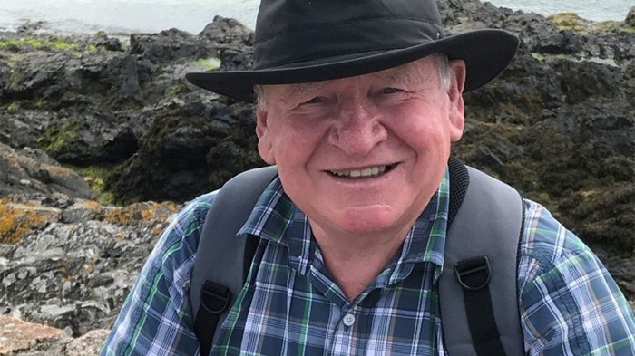 An older man - Gerald - in the countryside. He wears a brimmed hat, check shirt and has a backpack. The image is closely cropped with Gerald looking direct to camera and smiling.