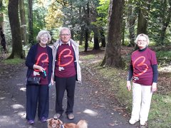 Three people wearing Blood Cancer UK t-shirts walk their dog through woodland, smiling at the camera