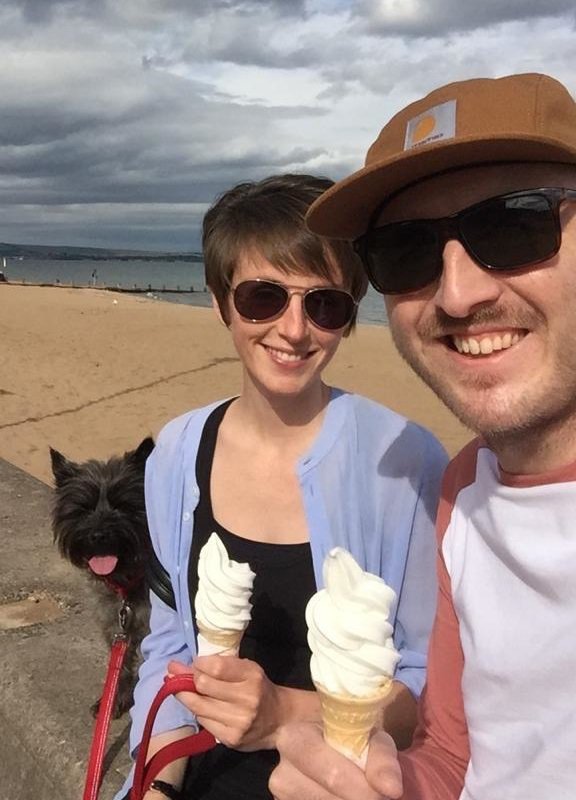 A selfie of Graeme and his wife on the beach, sitting together on a wall with the sand and sea behind them. They sit close together, smiling, eating ice creams, and their small black dog is sitting on the wall next to them also facing the camera.