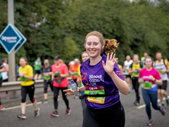 A woman in a purple Blood Cancer UK T shirt waves as she runs in a marathon.