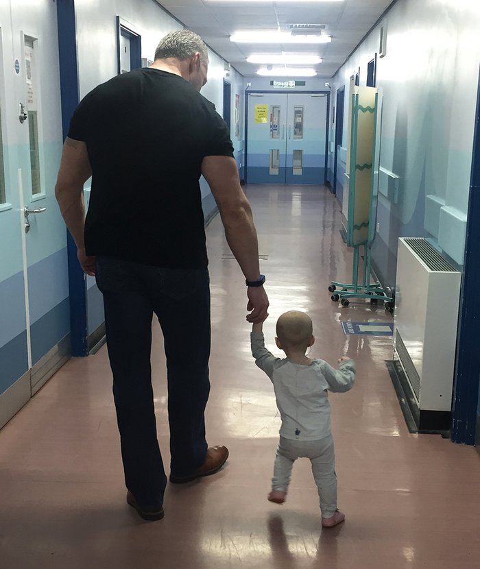 A very young child walks through a hospital corridor with her father.