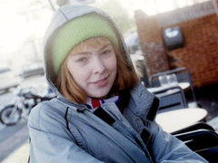 A woman in her 20s poses for a photograph outside. She's wearing a wooly hat and coat and sits at a cafe.