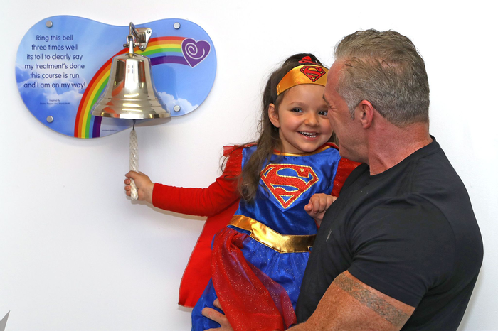 A young child Mia rings the end of treatment bell, held up by her father.