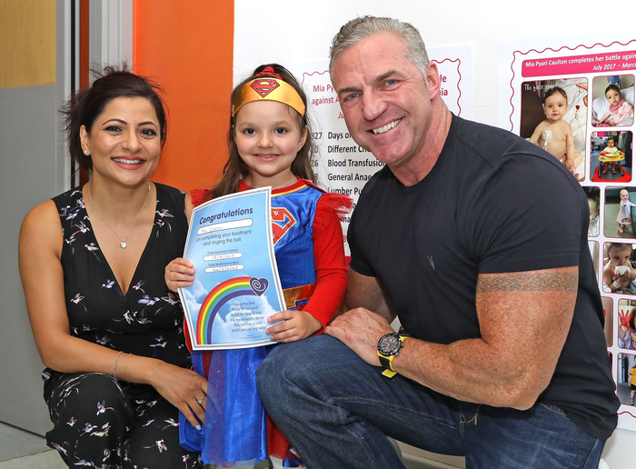 A young child - Mia - poses with a certificate dress as a superhero and flanked by two adults, a man and a woman.