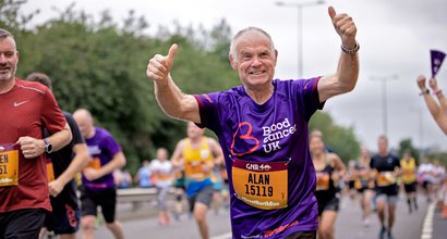 An older man in a purple Blood Cancer UK t-shirt holds his thumbs up as he runs a marathon.