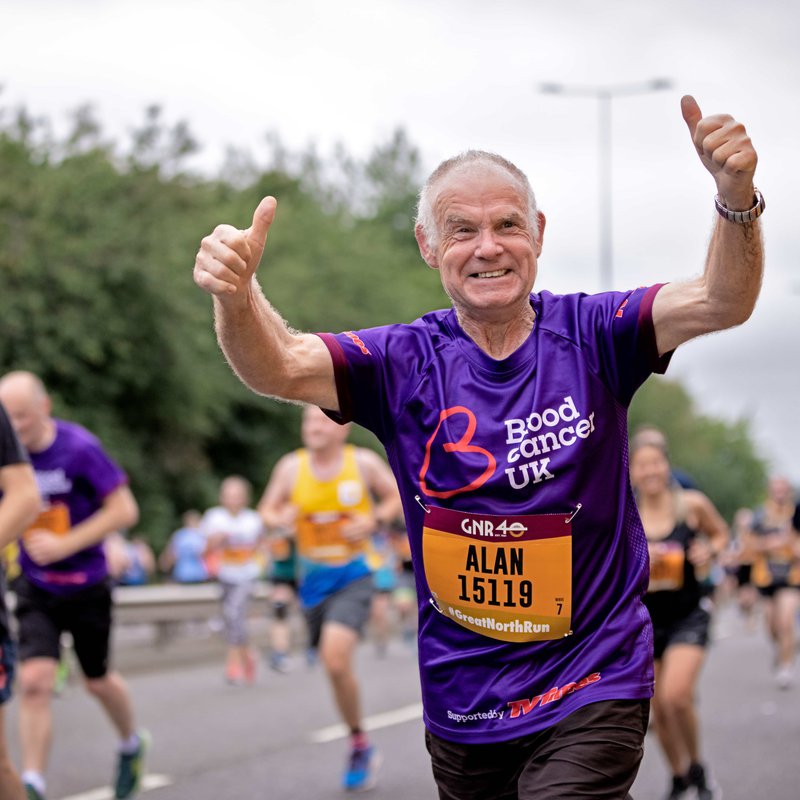 An older man in a purple Blood Cancer UK t-shirt holds his thumbs up as he runs a marathon.