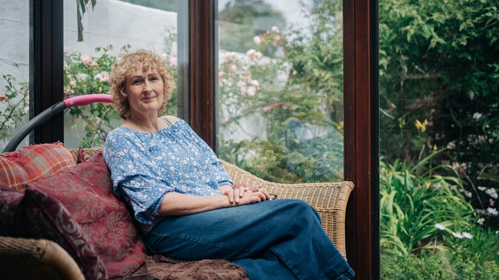 Rebecca sits in a cosy chair in her conservatory on a sunny day, smiling and relaxed.