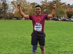 A runner celebrates after a marathon, arms held high in celebration, wearing a Blood Cancer UK T shirt in a park.