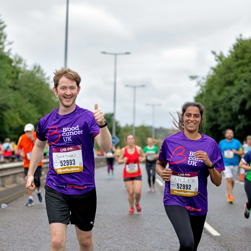 Two runners - a man and woman - smile as they run together wearing Blood Cancer UK t shirts during a marathon.