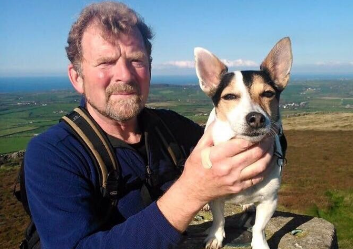 A man - Thomas - holds a pointy-eared dog as they pose on a country hillside