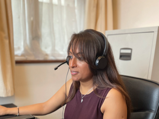A woman takes a call on a headset as she works from home, looking at the computer in front of her. She's seated at a leather office chair with a filing cabinet and window behind her.
