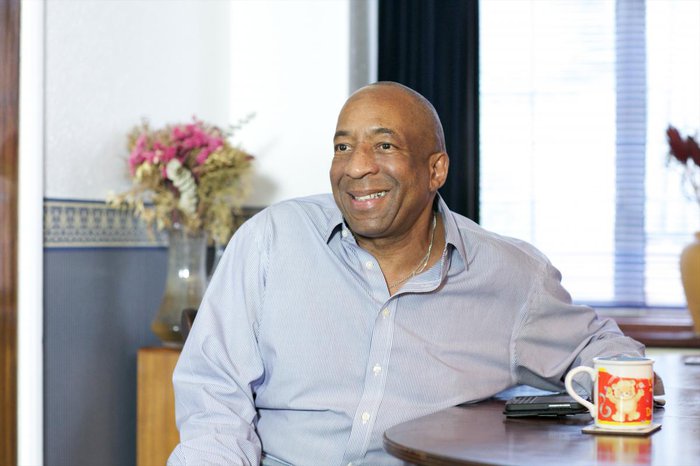 A dark-skinned, middle-aged man Newton smiles at home, with a cup on the table next to him.